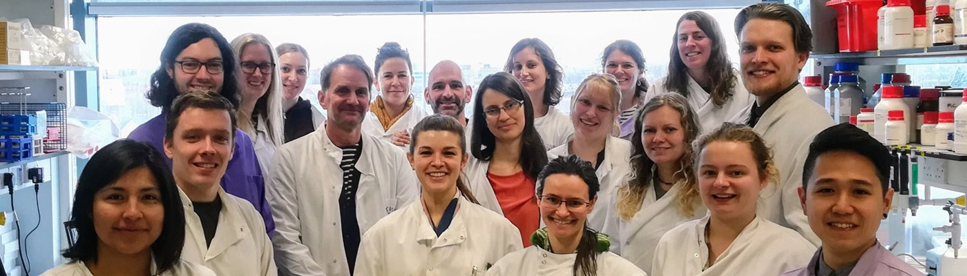 A group of scientists in white lab coats stand in a lab and smile for a group photo.