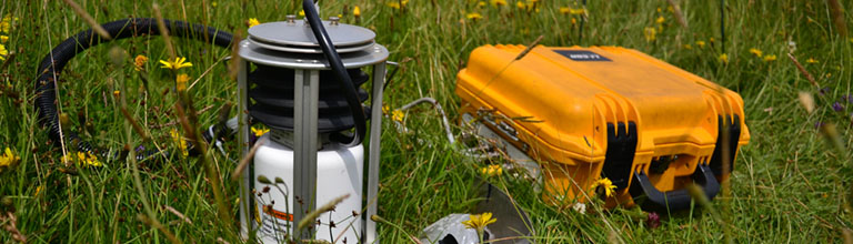 Scientific equipment and a yellow box lay among long grass in a meadow.